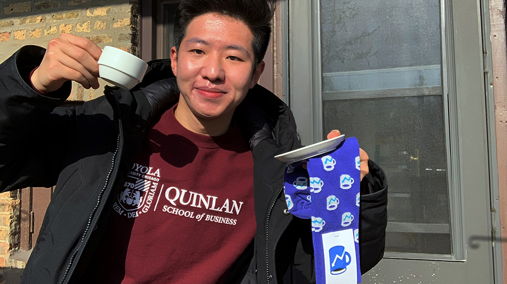 Student Jie Zheng smiling in a Quinlan t shirt, holding Morning Brew socks outside.
